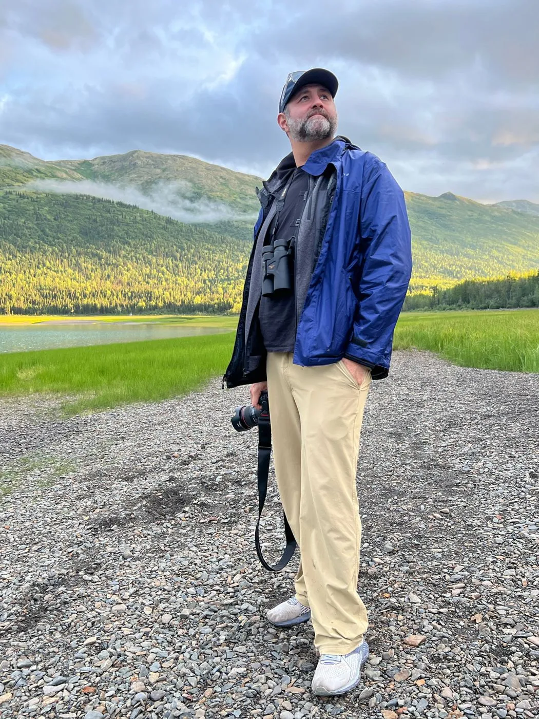 Temu Nana standing on a gravel shore with a camera, green hills behind
