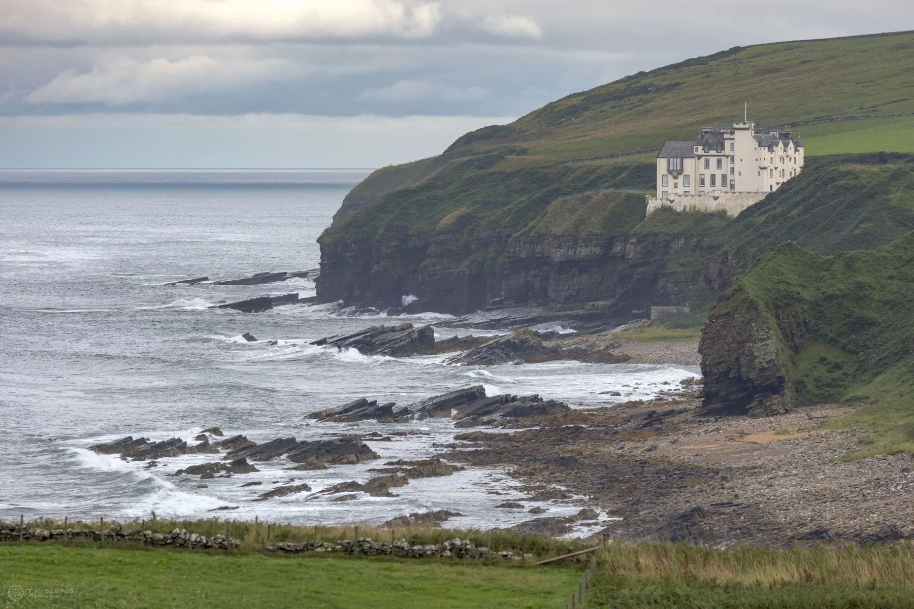 A white castle on a green headland above a rocky Scottish shore